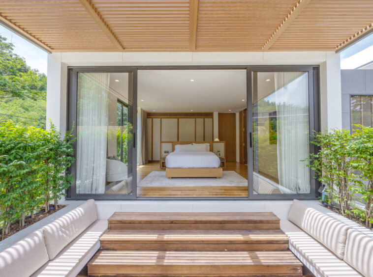 Bright modern bedroom seen through large glass sliding doors, with a low wooden bed, white bedding, and minimalist headboard, opening onto a wooden terrace framed by greenery outside.
