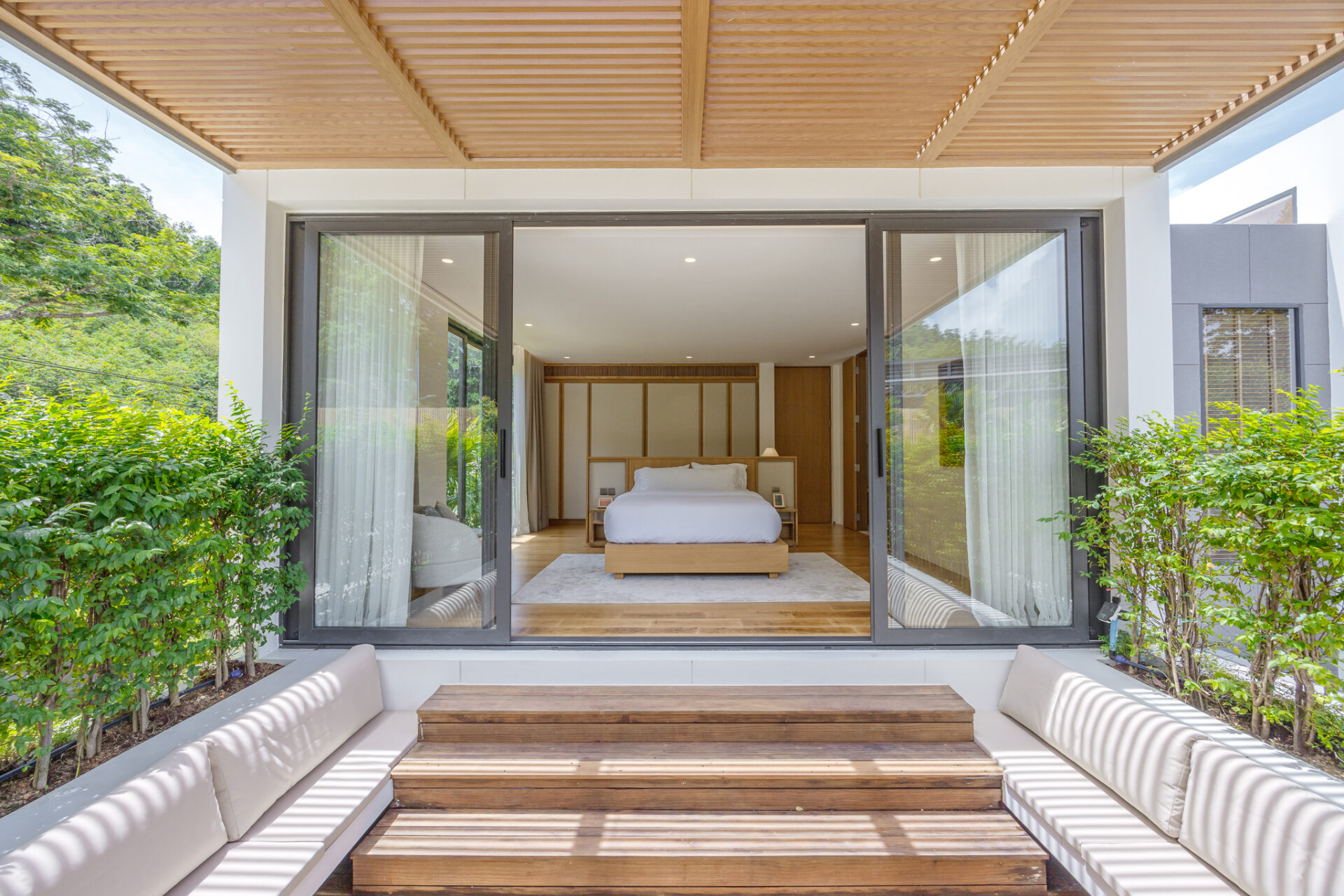Bright modern bedroom seen through large glass sliding doors, with a low wooden bed, white bedding, and minimalist headboard, opening onto a wooden terrace framed by greenery outside.