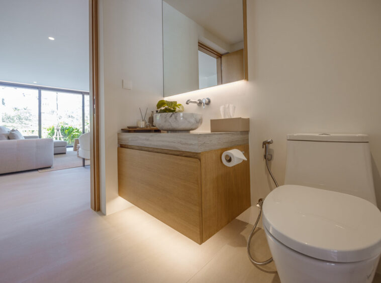 Modern bathroom with a wooden vanity, marble vessel sink, backlit mirror, and a white toilet; living room visible through the doorway.