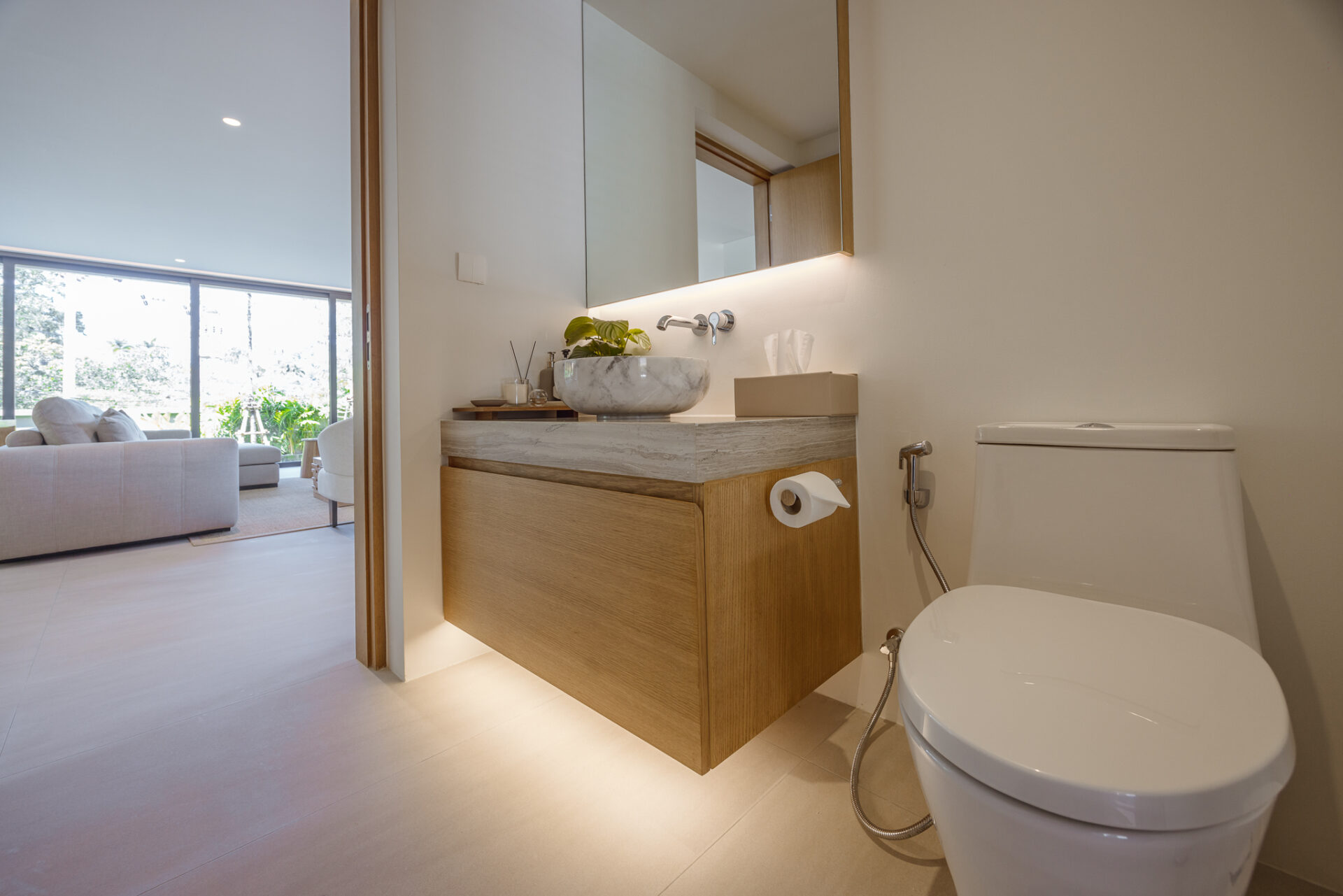Modern bathroom with a wooden vanity, marble vessel sink, backlit mirror, and a white toilet; living room visible through the doorway.