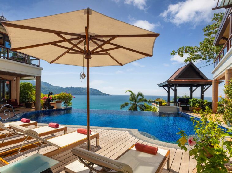 Resort pool deck with wooden lounge chairs and red towels, large beige umbrella, overlooking a calm blue ocean and distant green hills under a clear sky.