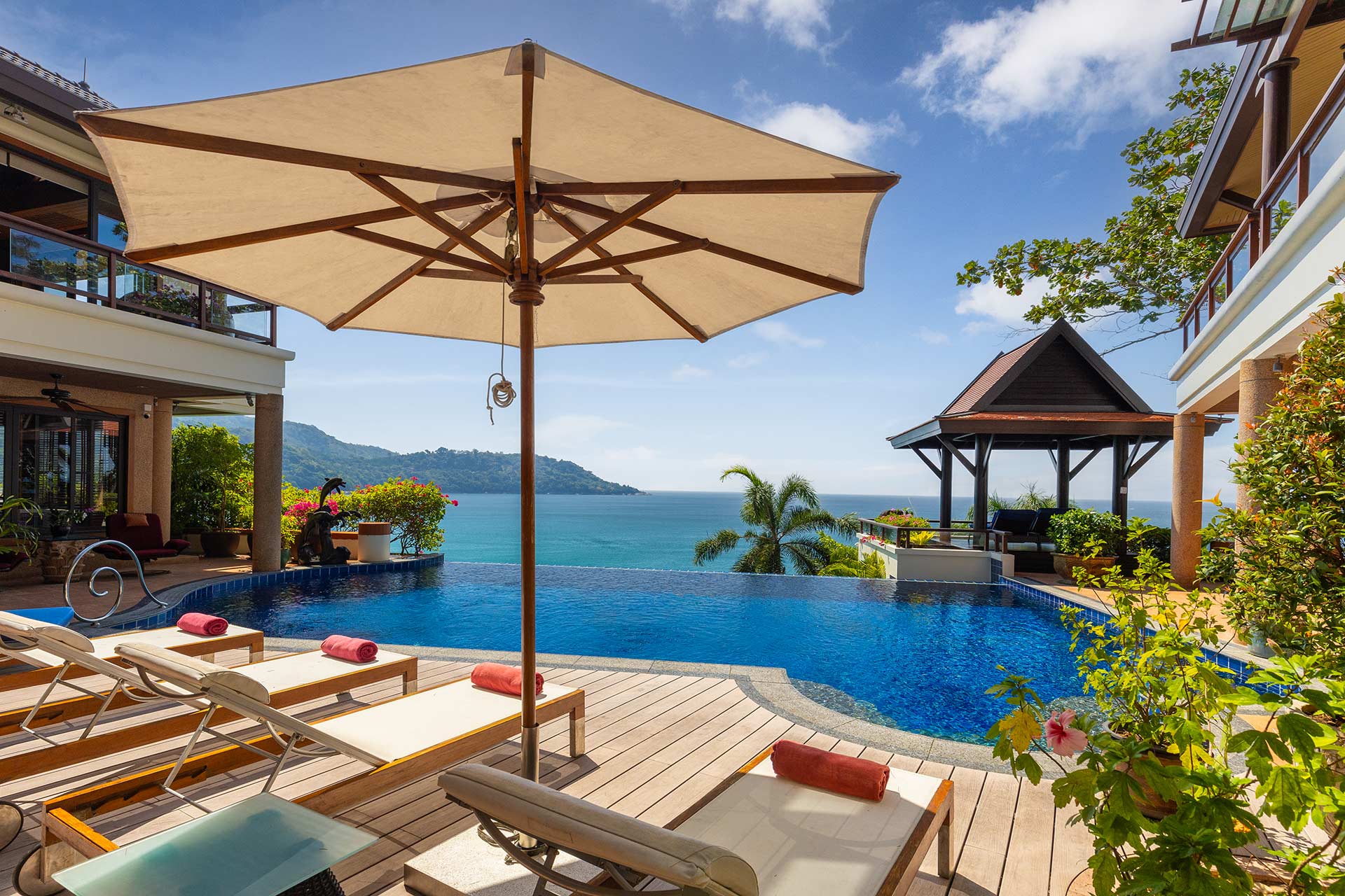 Resort pool deck with wooden lounge chairs and red towels, large beige umbrella, overlooking a calm blue ocean and distant green hills under a clear sky.