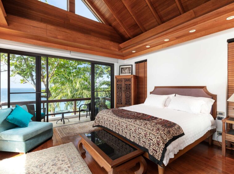 Hotel-style bedroom with a wooden beam ceiling and large ocean-view balcony doors; bed with patterned quilt and white linens, turquoise sofa nearby.