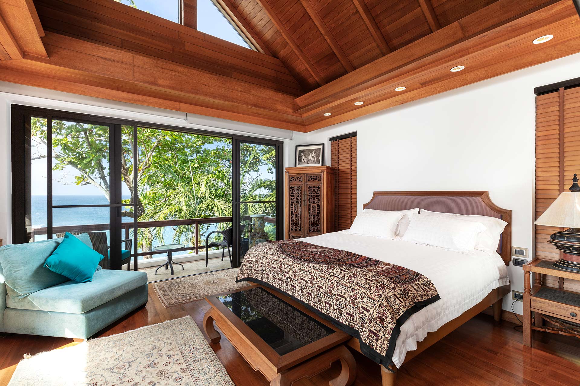 Hotel-style bedroom with a wooden beam ceiling and large ocean-view balcony doors; bed with patterned quilt and white linens, turquoise sofa nearby.