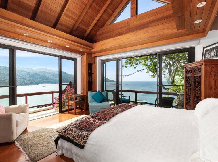 Oceanfront bedroom with a vaulted wooden ceiling, large glass doors, and a carved wooden armoire; view of the sea and hills beyond.