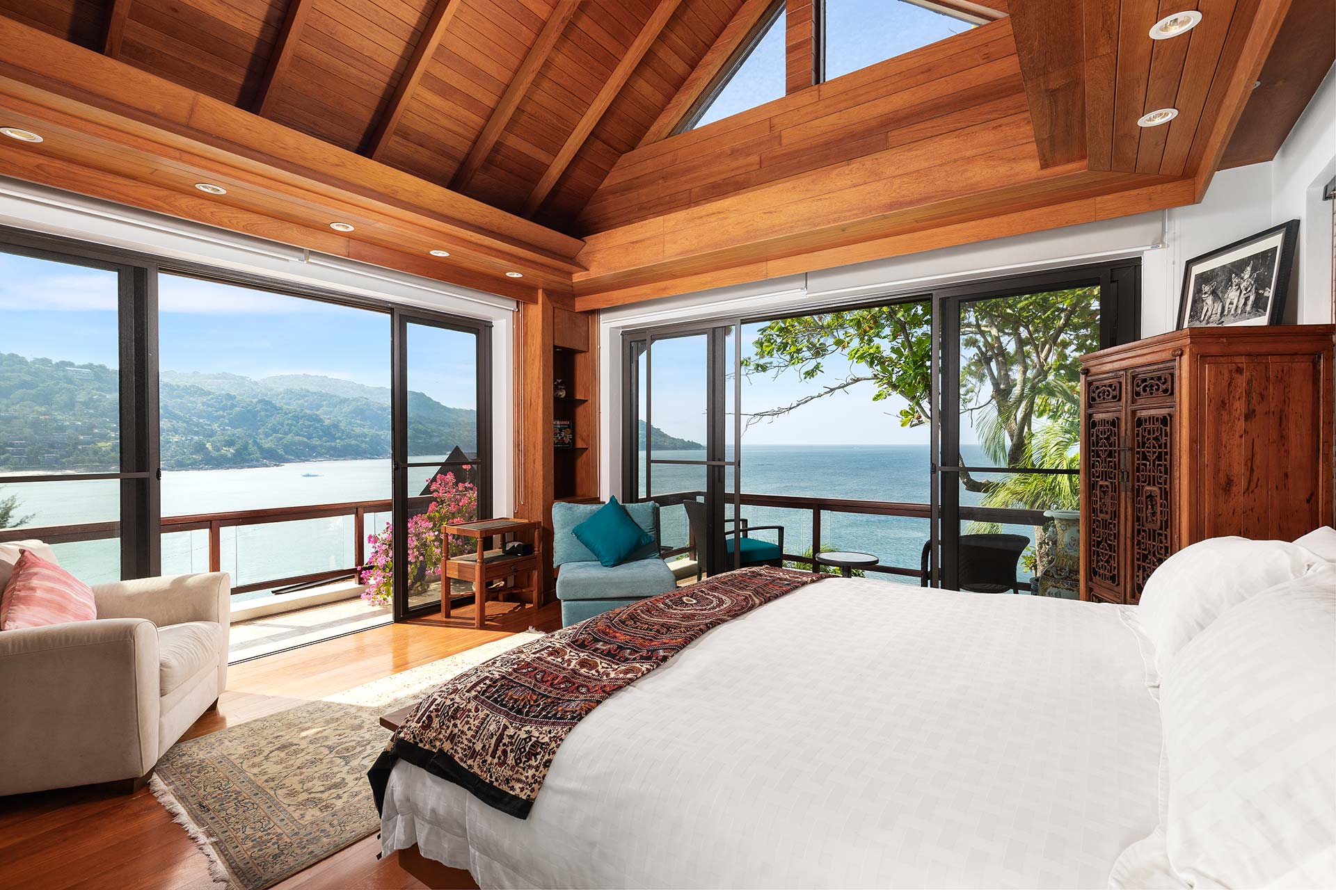 Oceanfront bedroom with a vaulted wooden ceiling, large glass doors, and a carved wooden armoire; view of the sea and hills beyond.