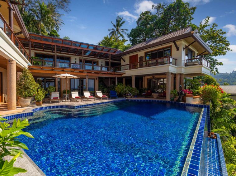 Resort-style villa with a large blue swimming pool, lounge chairs, and a covered patio overlooking tropical greenery and trees.