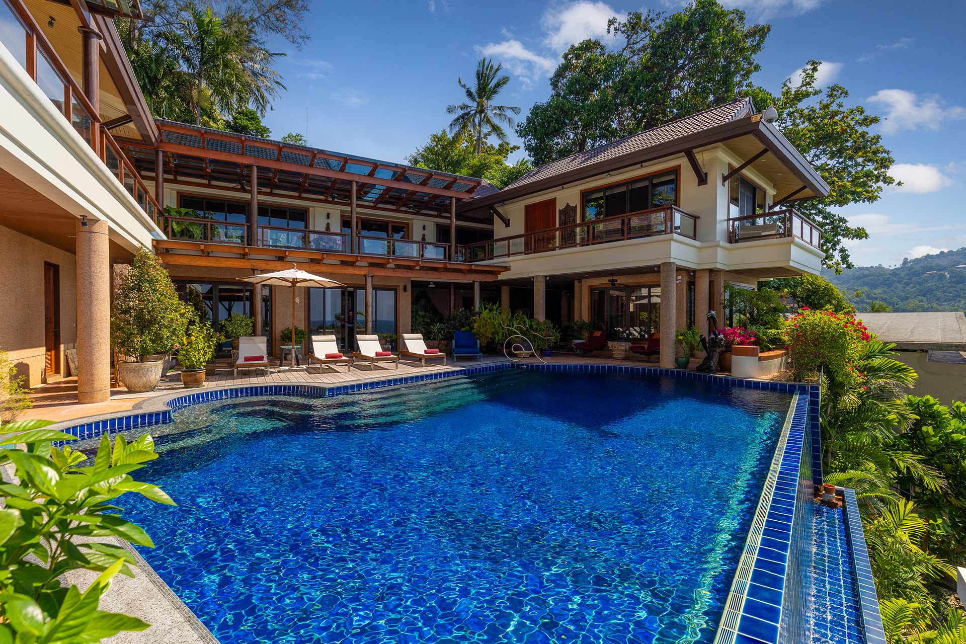 Resort-style villa with a large blue swimming pool, lounge chairs, and a covered patio overlooking tropical greenery and trees.