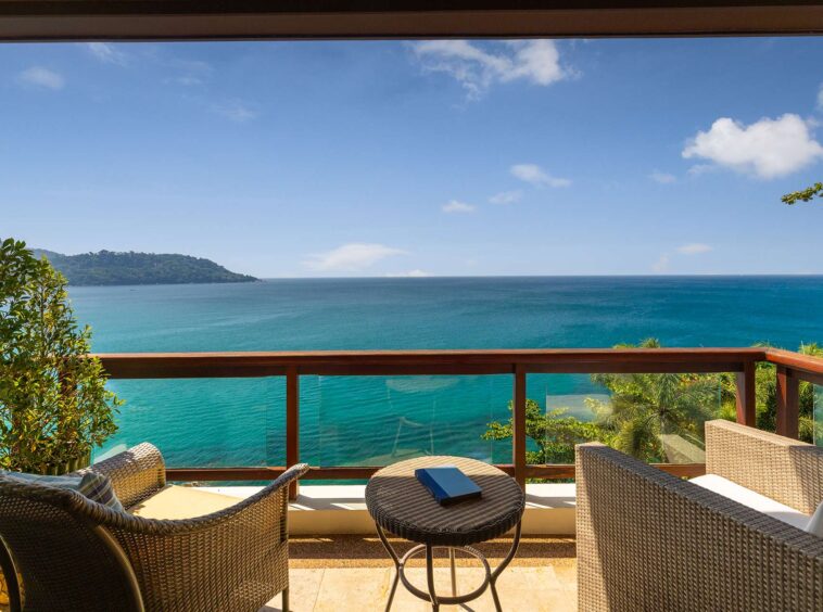 Balcony seating with wicker chairs and a glass railing overlooking turquoise sea and a distant island under a blue sky.