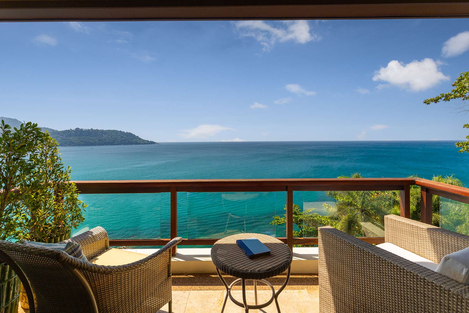 Balcony seating with wicker chairs and a glass railing overlooking turquoise sea and a distant island under a blue sky.