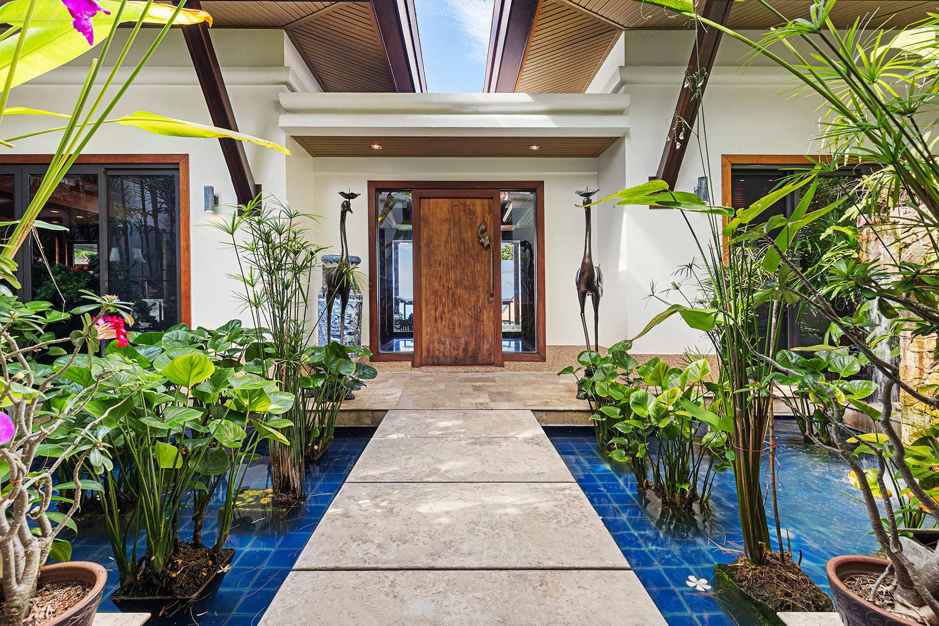 Entry courtyard with a wooden front door, glass sidelights, and lush tropical plants surrounding a blue-tiled water feature.