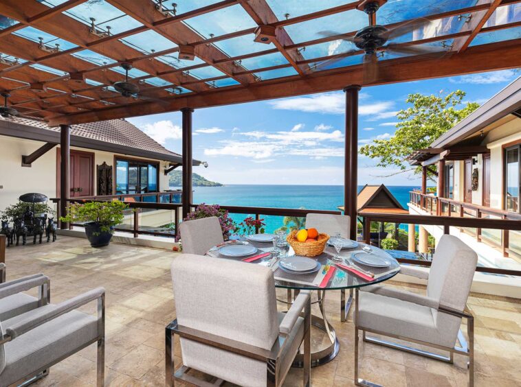 Luxury seaside terrace with a glass dining table set for six, fruit basket and tableware, overlooking the ocean and blue sky.