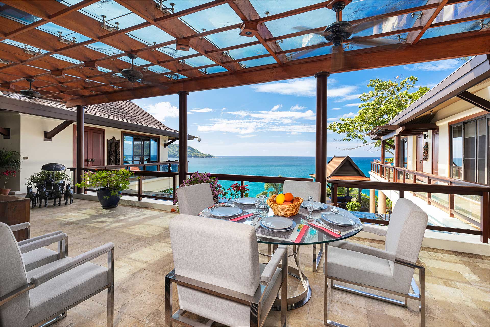 Luxury seaside terrace with a glass dining table set for six, fruit basket and tableware, overlooking the ocean and blue sky.