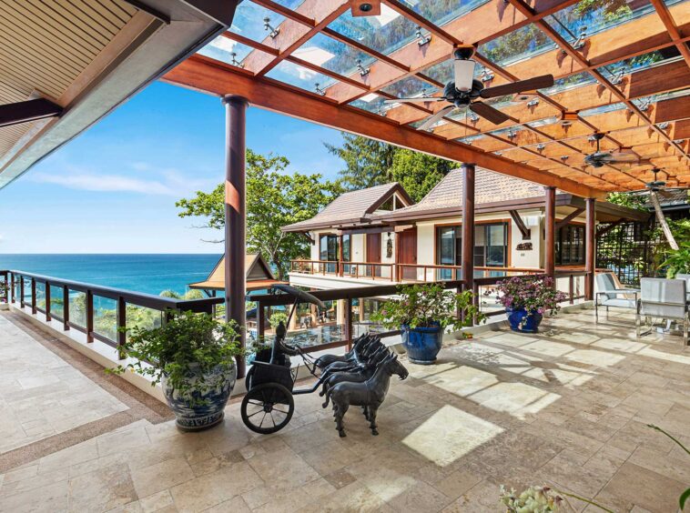 Seaside terrace with glass railing, beige tiled floor, potted plants, and a view of the ocean under a wooden pergola with ceiling fans.