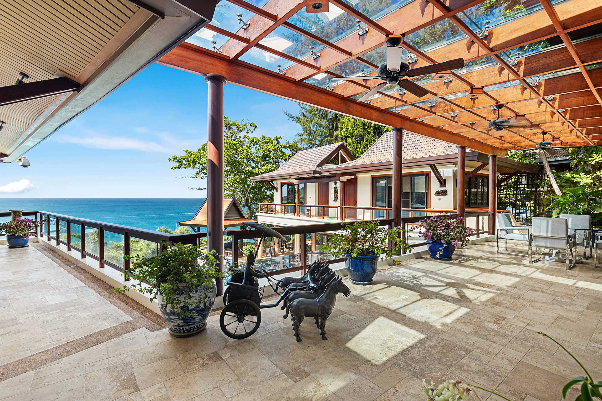 Seaside terrace with glass railing, beige tiled floor, potted plants, and a view of the ocean under a wooden pergola with ceiling fans.