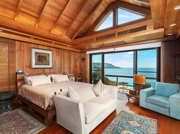 Wood-paneled bedroom with vaulted ceiling, beige bed, and ocean-view windows.