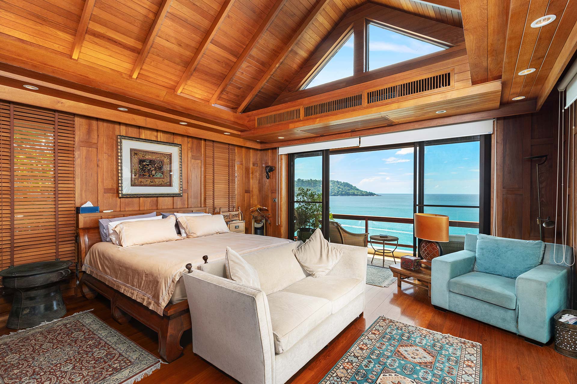 Wood-paneled bedroom with vaulted ceiling, beige bed, and ocean-view windows.