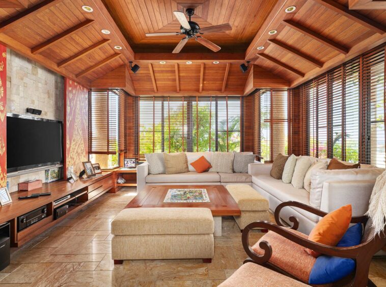 Living room with a warm wood ceiling, beige sectional sofa, and large windows with wooden blinds letting in sunlight.