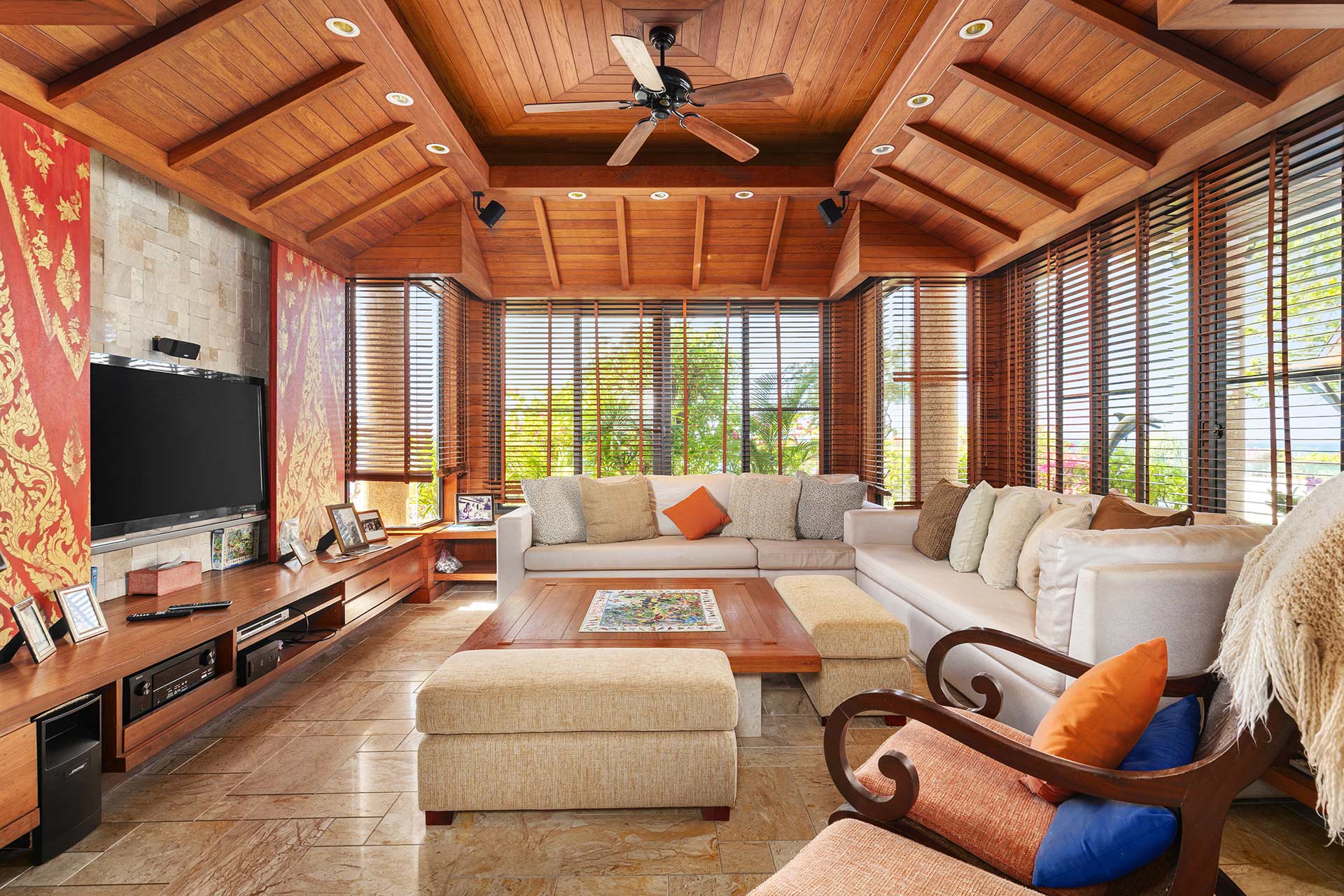 Living room with a warm wood ceiling, beige sectional sofa, and large windows with wooden blinds letting in sunlight.