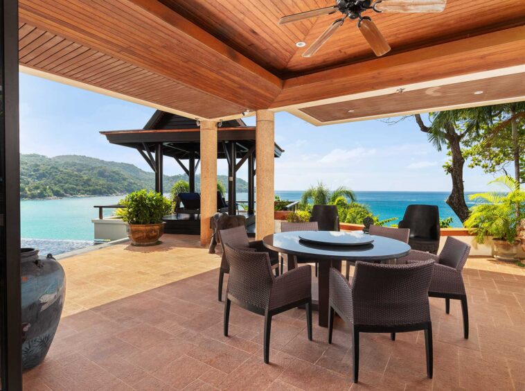 Outdoor patio with wicker chairs around a round table, overlooking the blue ocean and distant hills.