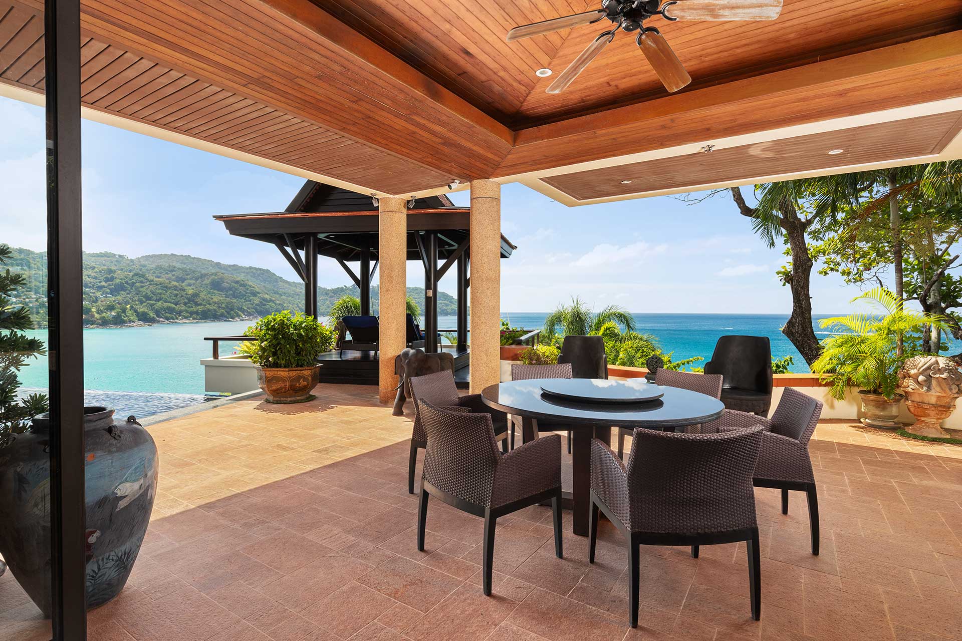 Outdoor patio with wicker chairs around a round table, overlooking the blue ocean and distant hills.