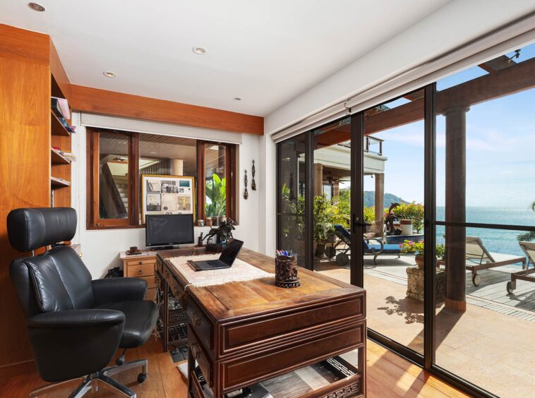 Bright home office with a dark wooden desk and black leather chair, facing large glass doors to a sea-view patio.