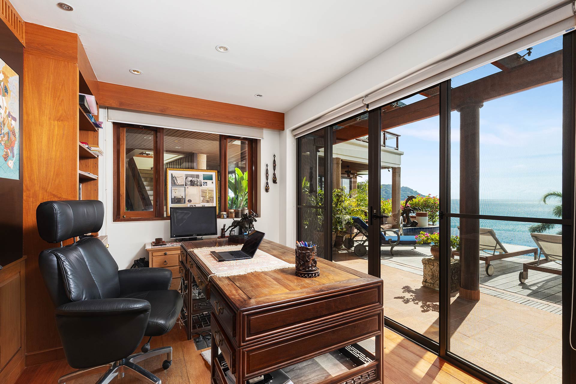 Bright home office with a dark wooden desk and black leather chair, facing large glass doors to a sea-view patio.