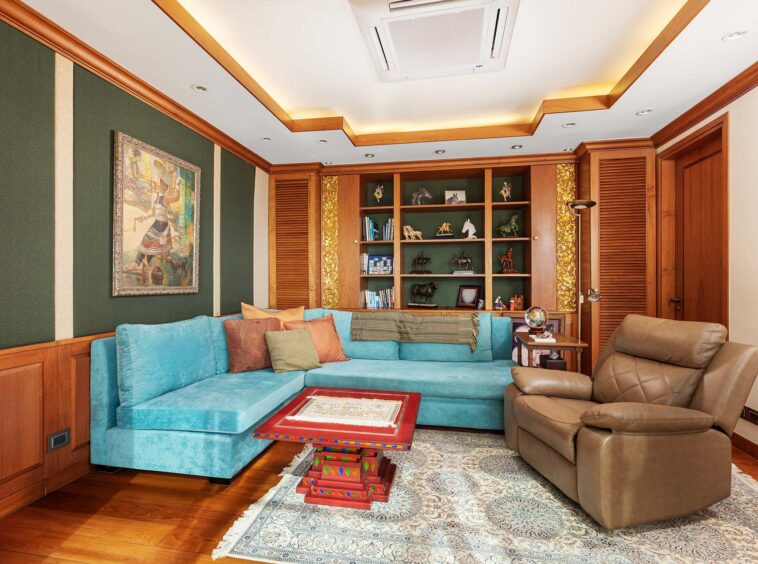 A bright living room with a teal sectional sofa, brown recliner, and a wooden built-in bookshelf filled with decorative items and books against green walls with wood trim.