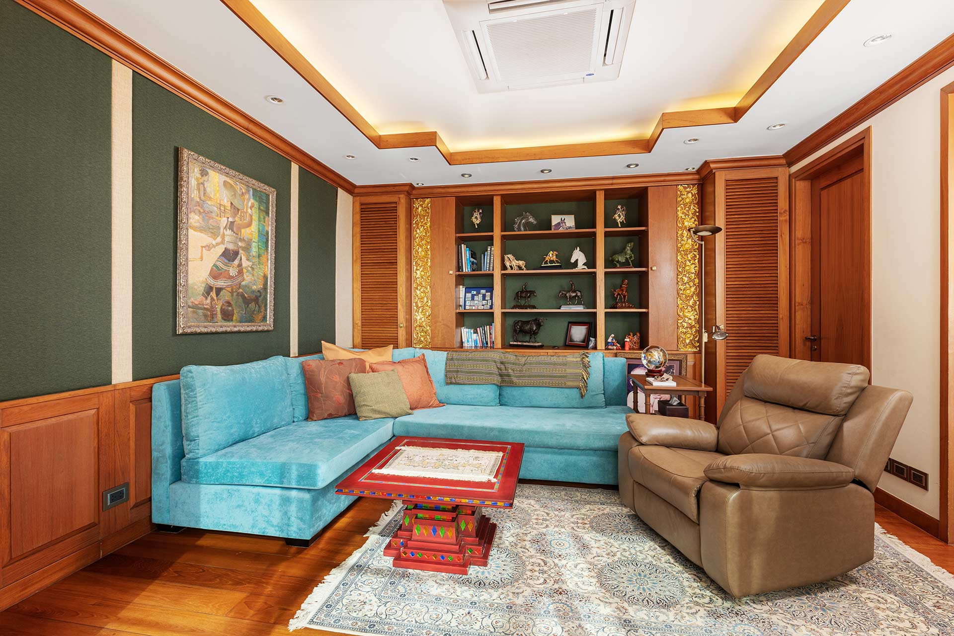 A bright living room with a teal sectional sofa, brown recliner, and a wooden built-in bookshelf filled with decorative items and books against green walls with wood trim.