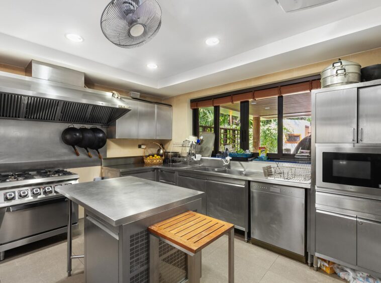 Commercial stainless-steel kitchen featuring a gas range with a large vent hood, hanging pots, and a window view to an outdoor patio garden. Ceiling fan and recessed lights visible.