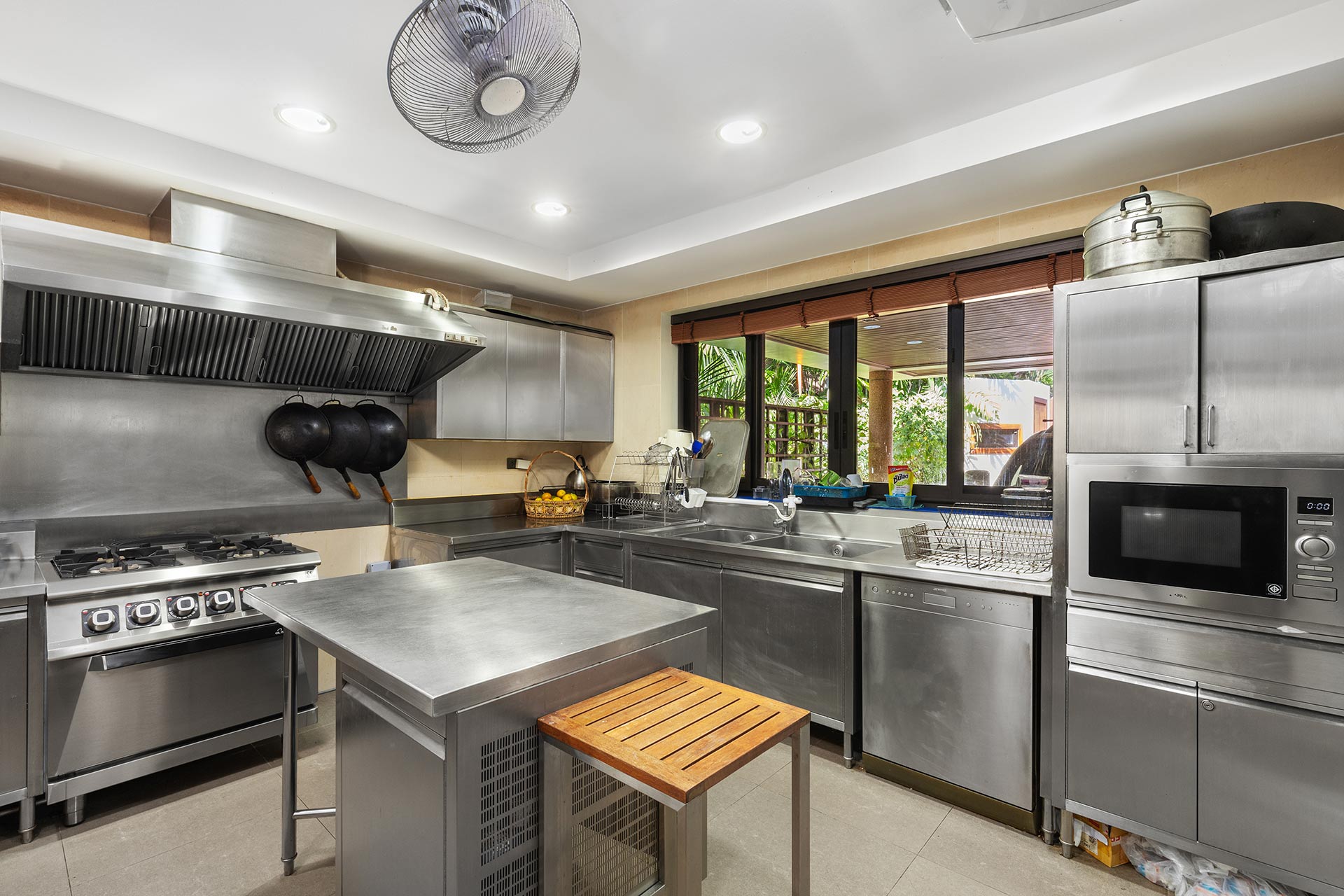 Commercial stainless-steel kitchen featuring a gas range with a large vent hood, hanging pots, and a window view to an outdoor patio garden. Ceiling fan and recessed lights visible.
