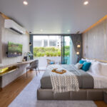 Modern hotel bedroom with a king bed, gray tones, teal accent wall, and wall-mounted TV overlooking a garden balcony.