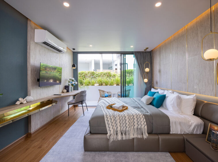 Modern hotel bedroom with a king bed, gray tones, teal accent wall, and wall-mounted TV overlooking a garden balcony.