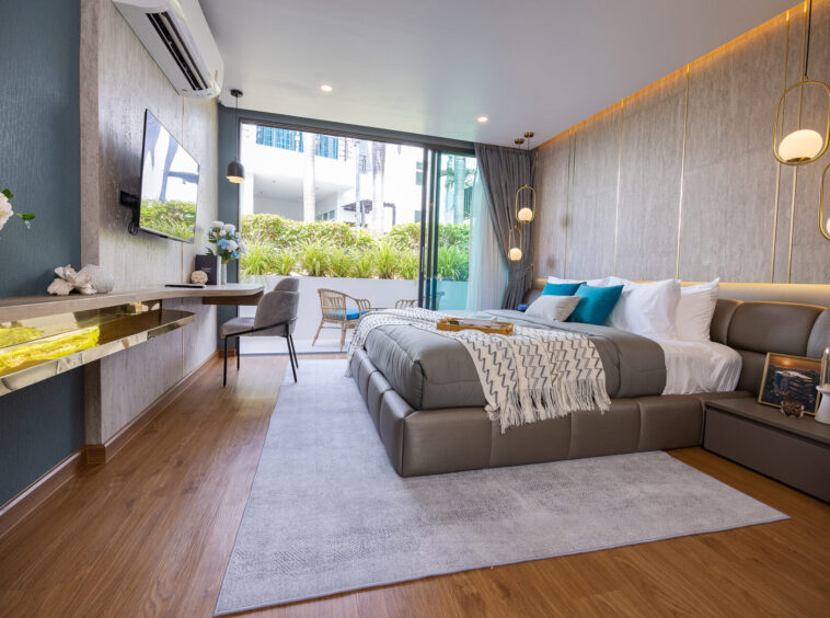 Modern hotel bedroom with a large gray bed, teal pillows, and a padded headboard; glass doors open to a balcony with greenery outside.