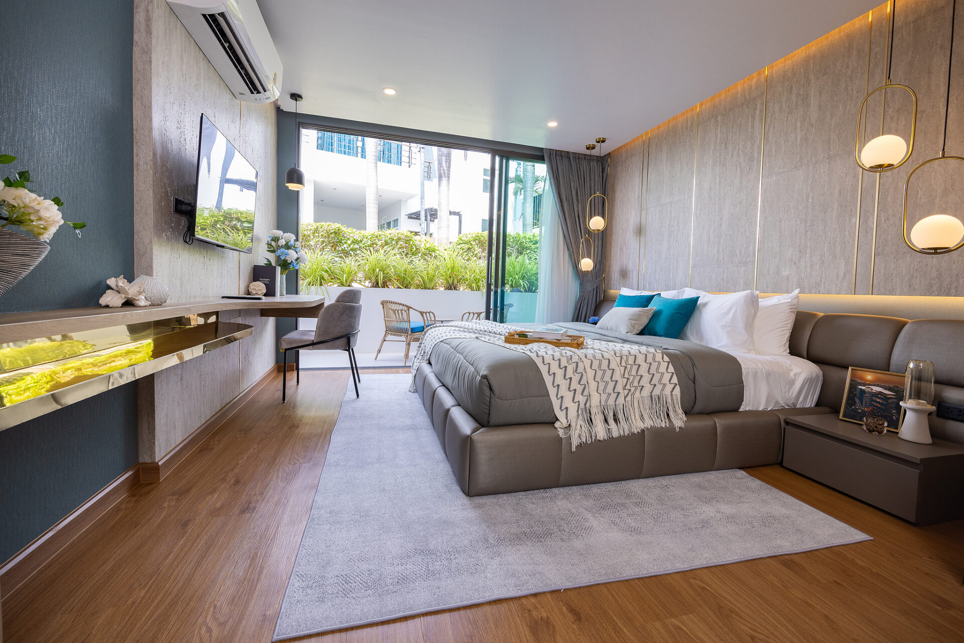 Modern hotel bedroom with a large gray bed, teal pillows, and a padded headboard; glass doors open to a balcony with greenery outside.