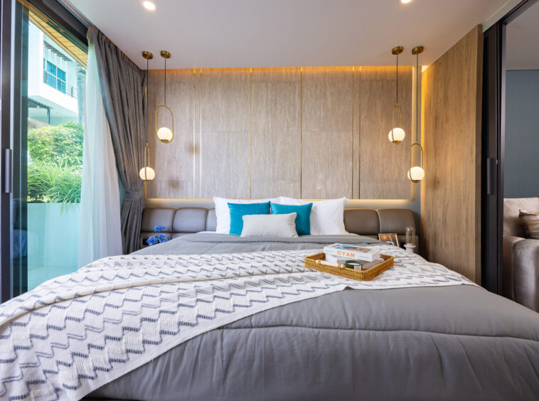 Modern hotel bedroom with a gray upholstered headboard, white and teal pillows, and a zigzag throw on a king bed.