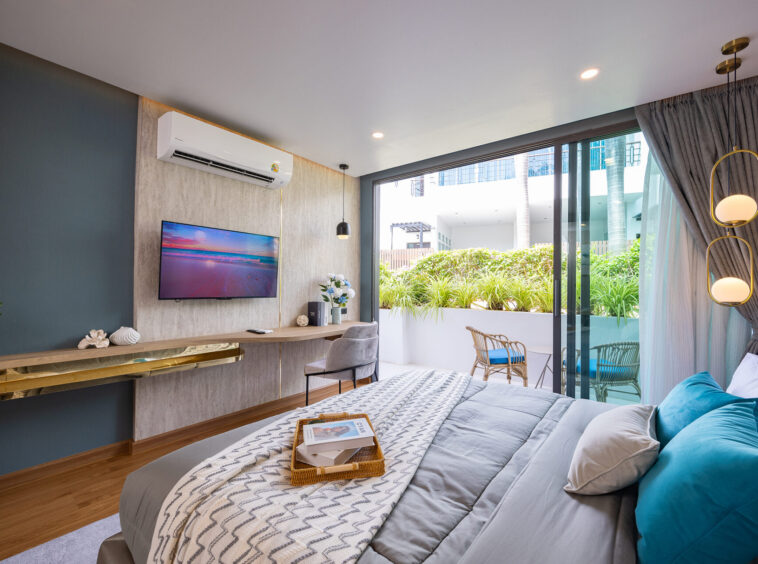 Modern hotel bedroom with a wall-mounted TV, air conditioner, and glass doors opening to a sunny balcony with plants outside the room.