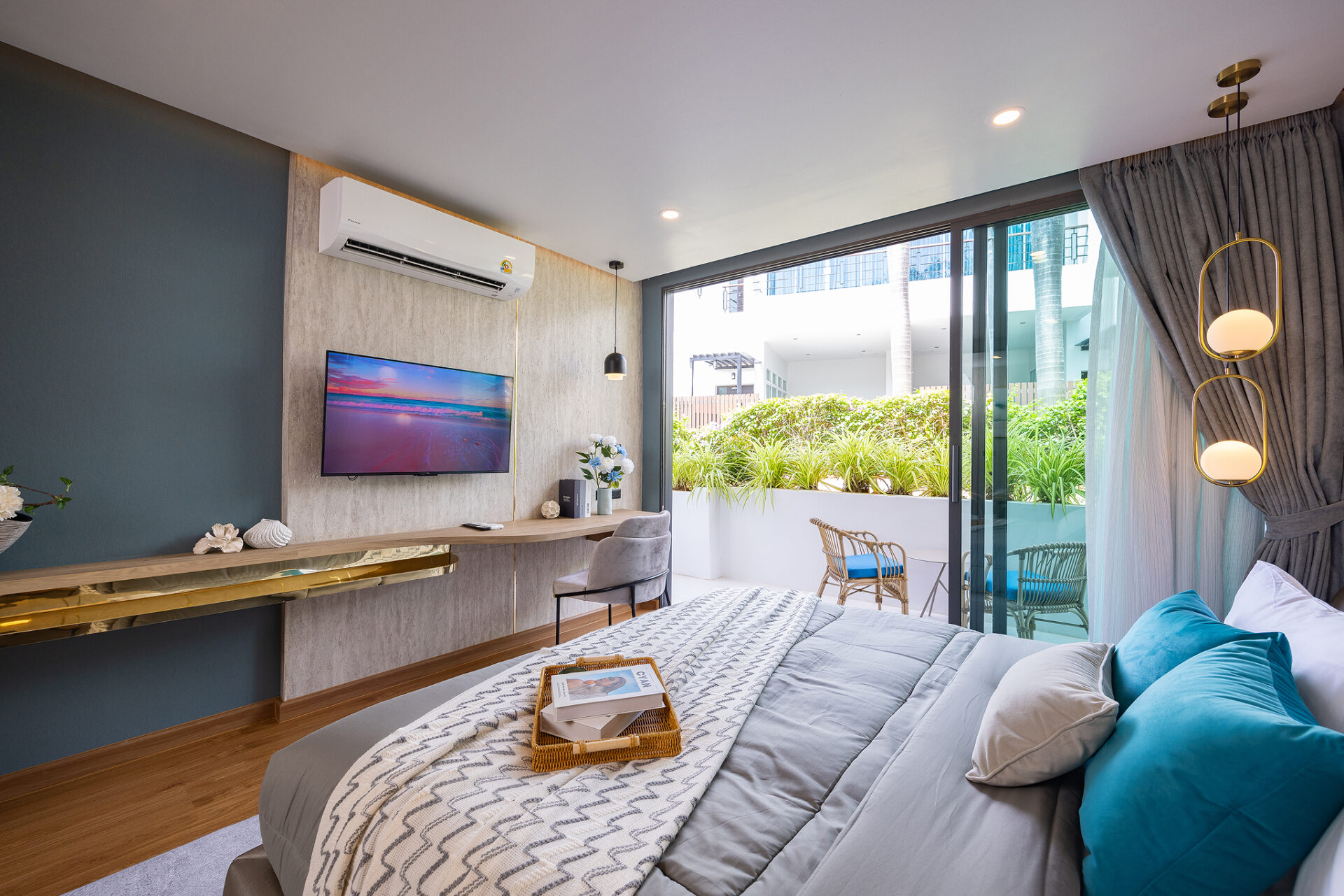 Modern hotel bedroom with a wall-mounted TV, air conditioner, and glass doors opening to a sunny balcony with plants outside the room.