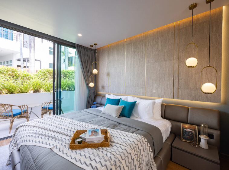 Modern hotel bedroom with a large bed, grey headboard, blue-accent pillows, and a woven tray with magazines on the duvet; glass doors open to a sunny balcony with wicker seating and greenery outside.