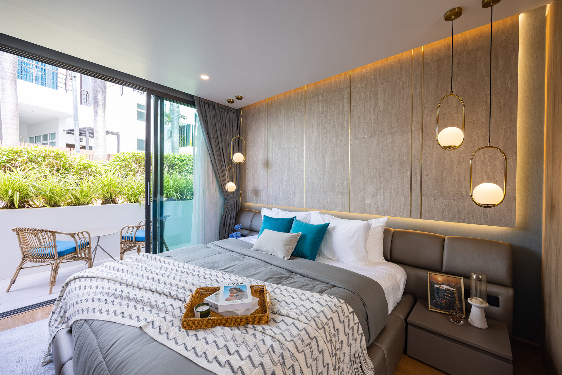 Modern hotel bedroom with a large bed, grey headboard, blue-accent pillows, and a woven tray with magazines on the duvet; glass doors open to a sunny balcony with wicker seating and greenery outside.