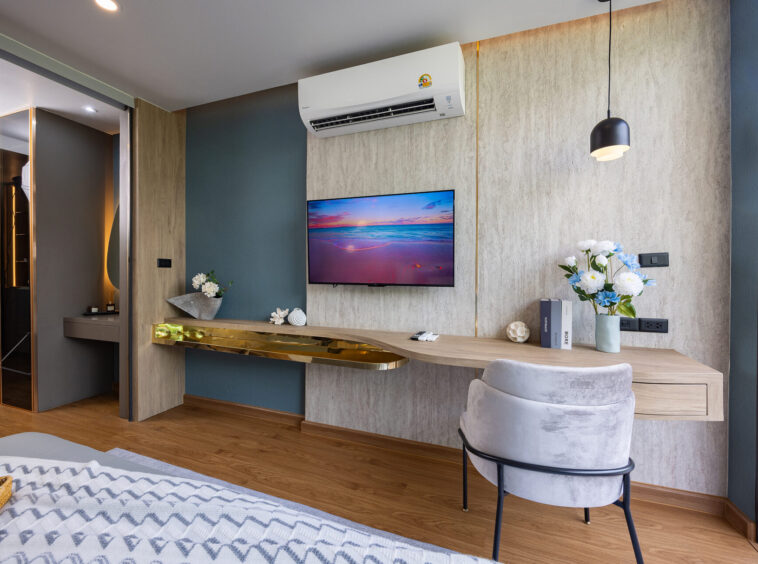 Modern hotel living area with a wall-mounted TV, air conditioner above, and a wooden desk with flowers below the TV.