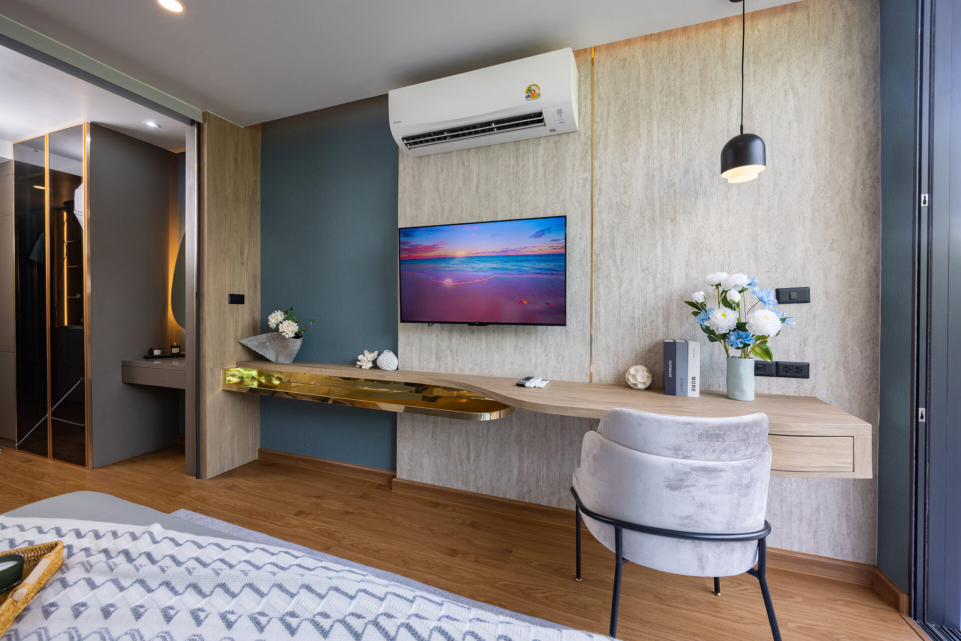 Modern hotel living area with a wall-mounted TV, air conditioner above, and a wooden desk with flowers below the TV.