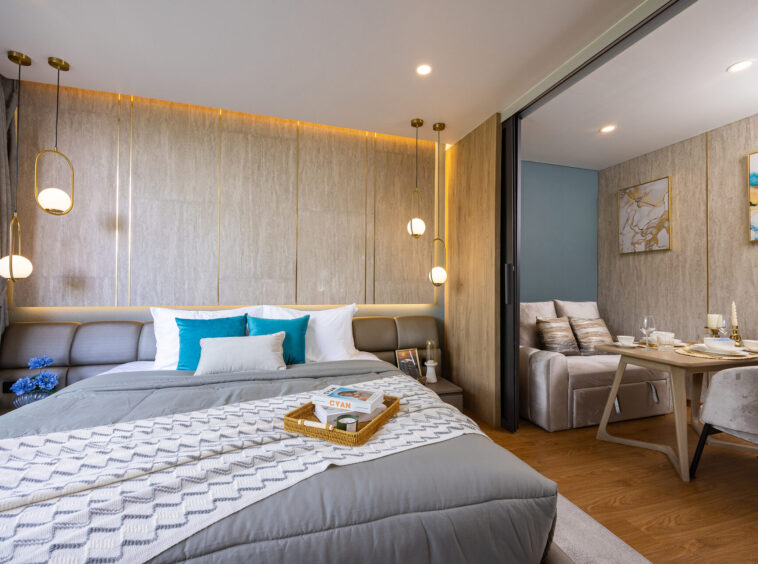 Modern hotel bedroom with a gray upholstered bed, blue accent pillows, and a wicker tray on a patterned duvet near a nightstand and hanging globe lights.