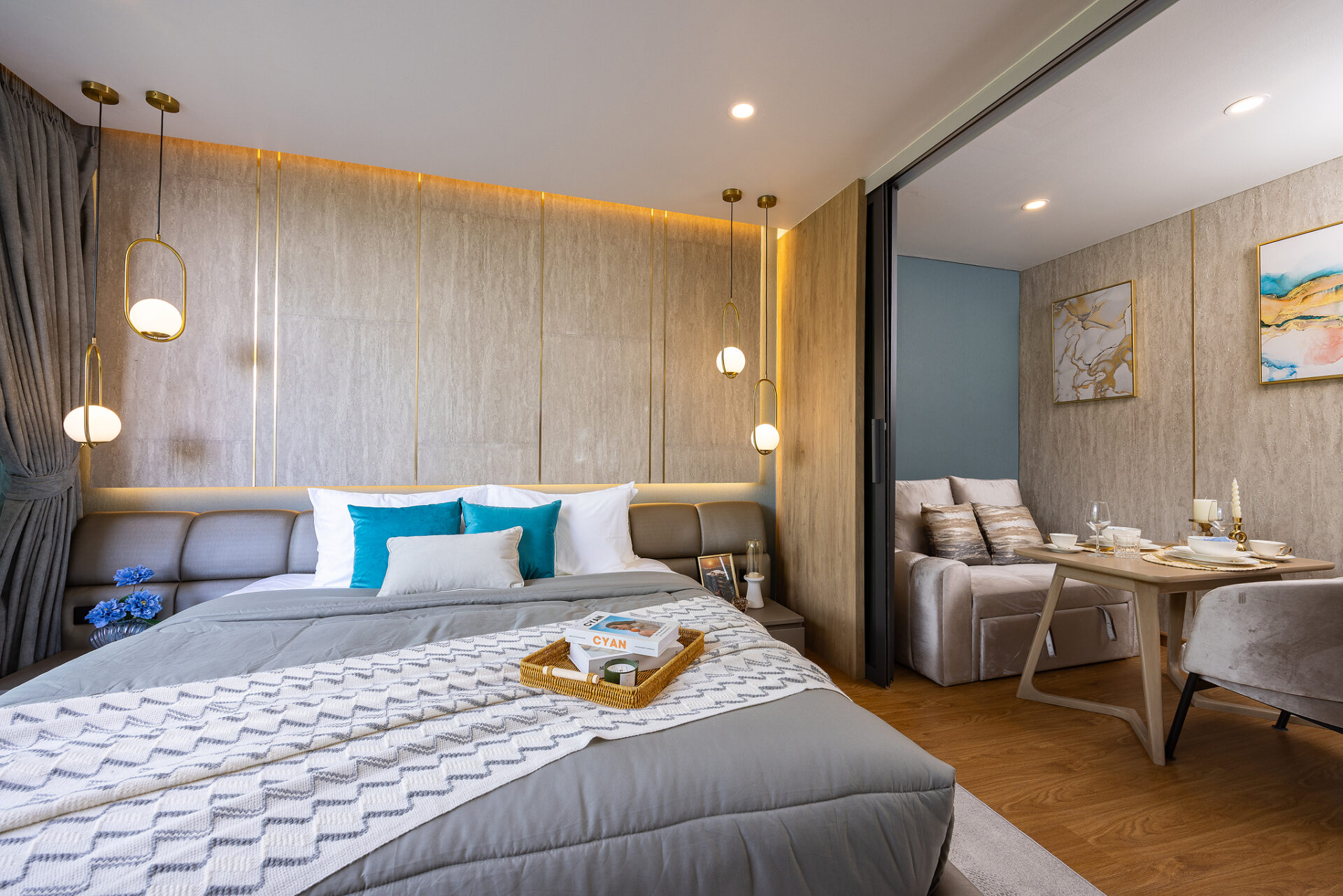 Modern hotel bedroom with a gray upholstered bed, blue accent pillows, and a wicker tray on a patterned duvet near a nightstand and hanging globe lights.