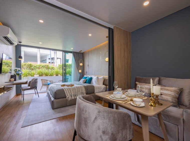 Open-plan hotel suite with dining area and gray sofas, separated from the bedroom by a sliding glass wall and outdoor greenery visible outside.