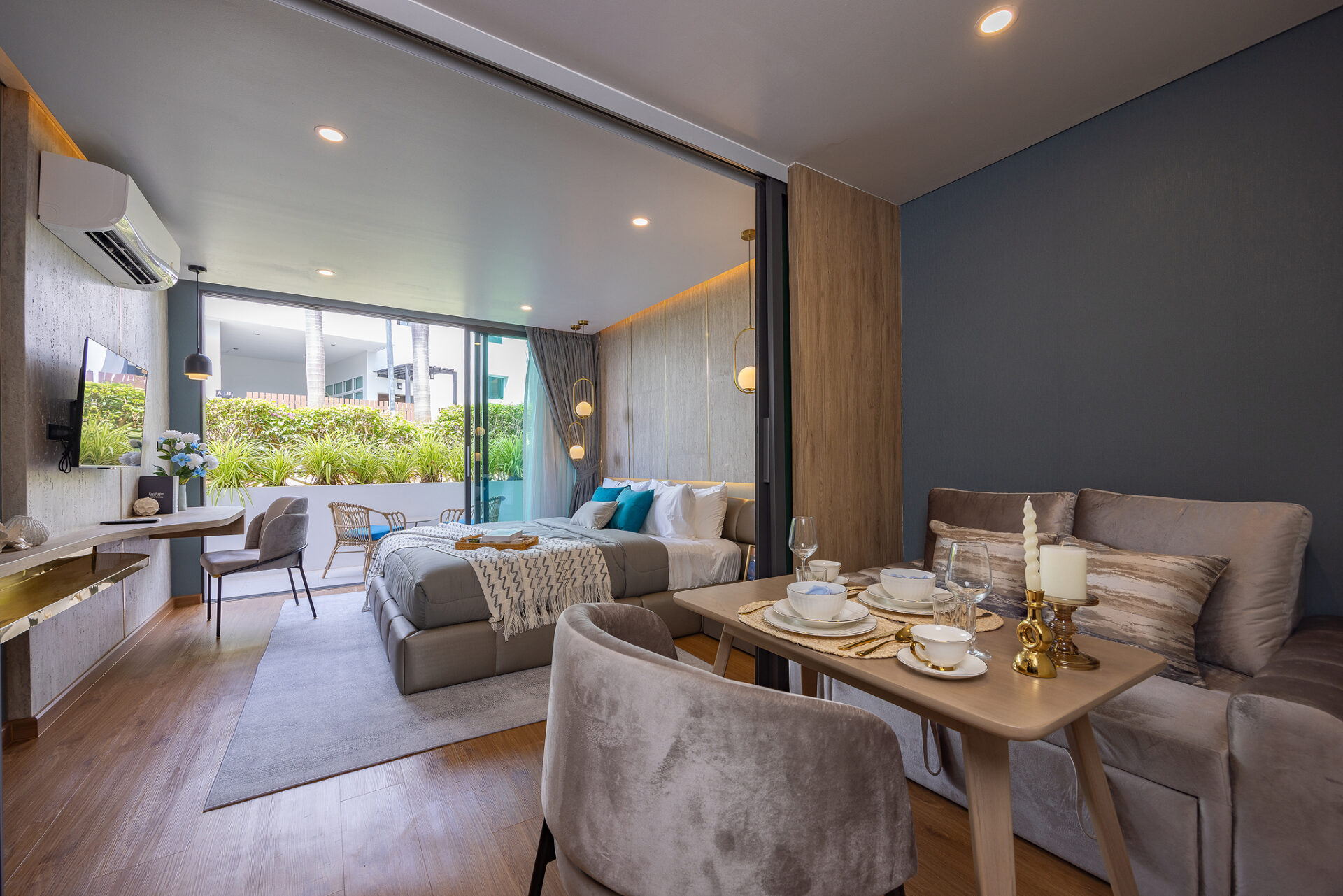 Open-plan hotel suite with dining area and gray sofas, separated from the bedroom by a sliding glass wall and outdoor greenery visible outside.