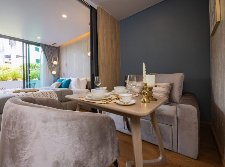 Modern living-dining space with a beige sectional, wooden table set for two, and gold candleholders near a glass door to a sunny balcony.