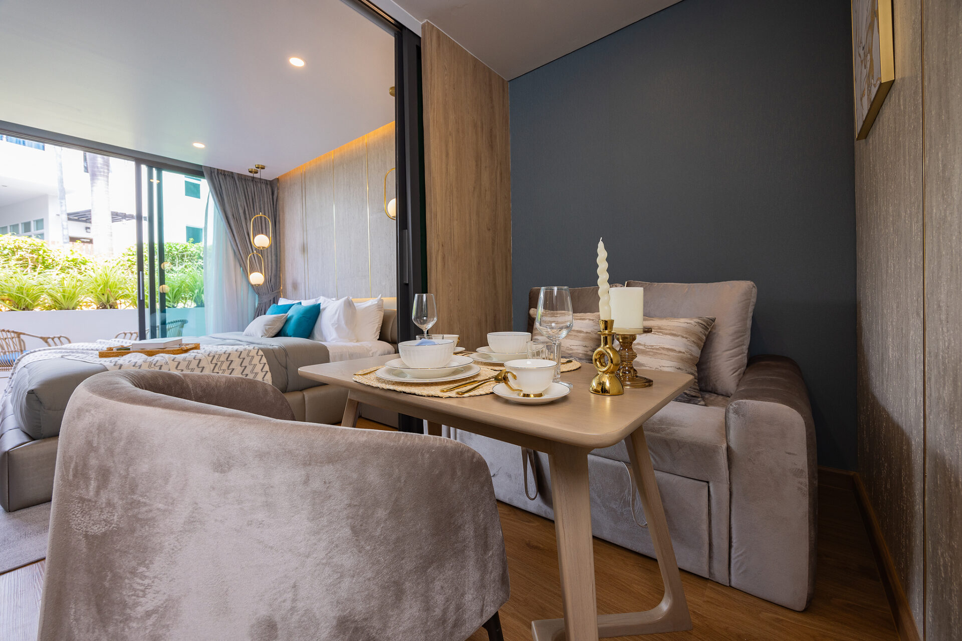Modern living-dining space with a beige sectional, wooden table set for two, and gold candleholders near a glass door to a sunny balcony.
