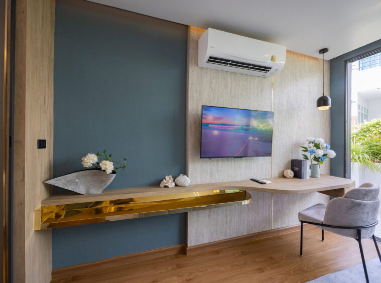 Modern living room with a wall-mounted TV, ceiling air conditioner, blue accent wall, wooden desk, and a gold decorative shelf along the wall.
