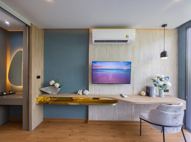 Modern living area with a wall-mounted TV, white air conditioner, blue and beige walls, and a wooden desk with a gold shelf.